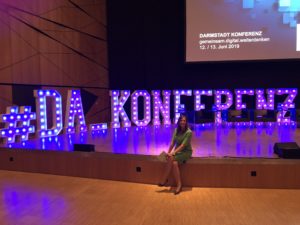 Moderatorin Kristina zur Mühlen sitzt auf Bühnenkante | blau und lila leuchtende Großbuchstaben auf Bühne | "#DA_Konferenz"